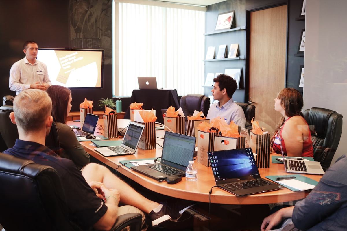 Un instructor exponiendo ante un grupo sentado alrededor de una mesa de juntas con sus computadoras abiertas.